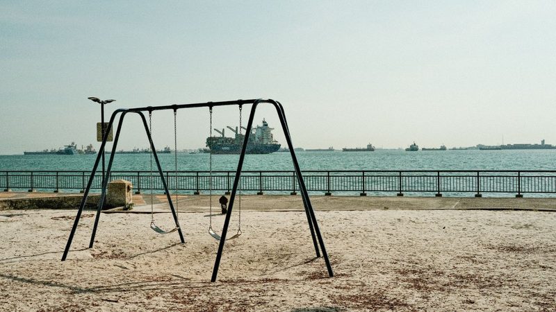 Tranquil playground with swings by Singapore's waterfront, ships in the background.