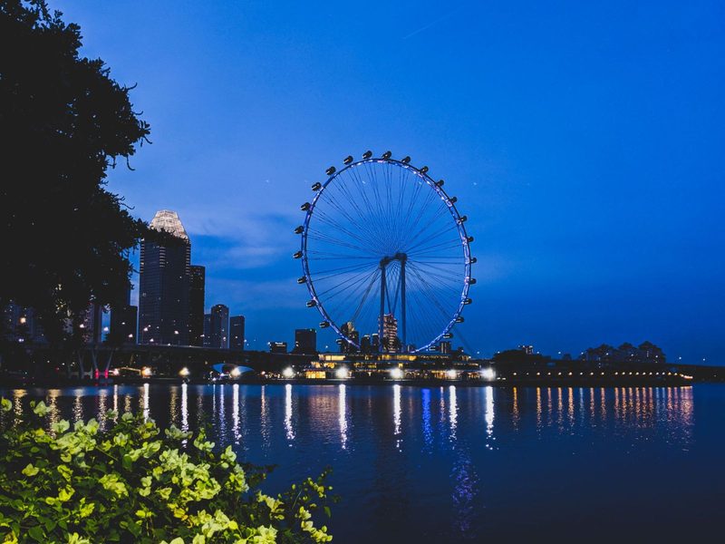 Scenic view of the illuminated Singapore Flyer against a city skyline at dusk, reflecting on water.