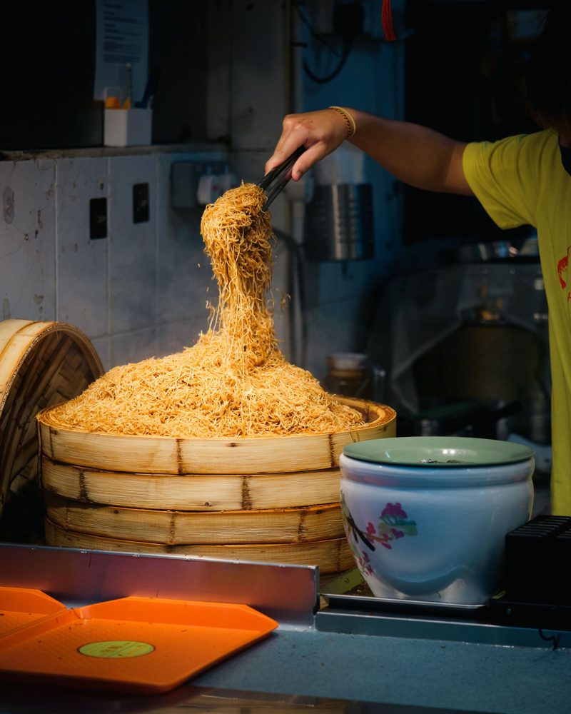 A street vendor serving a large bamboo steamer filled with Asian noodles in a bustling market.