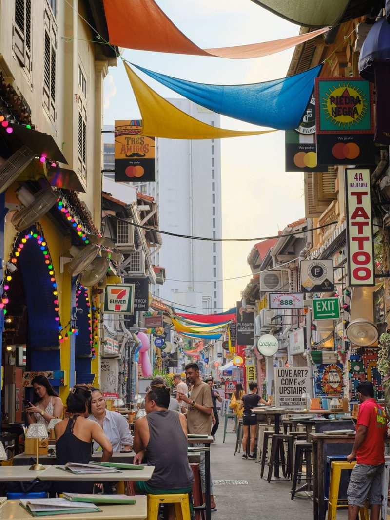 Vibrant street view of Haji Lane in Singapore filled with colorful banners, shops, and people socializing outdoors.