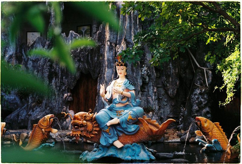 Colorful Bodhisattva sculpture on a mythological creature surrounded by greenery in a tranquil garden setting.