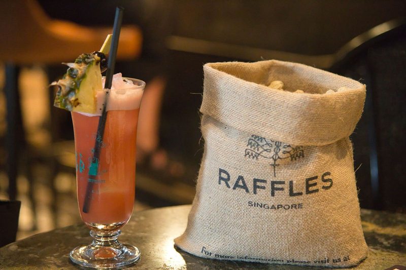Glass of Singapore Sling with pineapple garnish beside a Raffles bag.