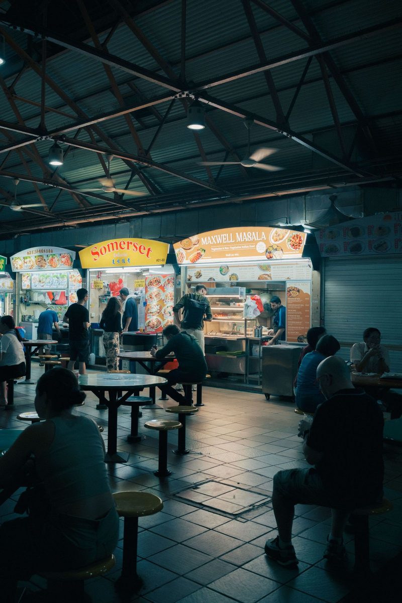 A vibrant night scene at Maxwell Food Centre, showcasing diverse food stalls and seated patrons.